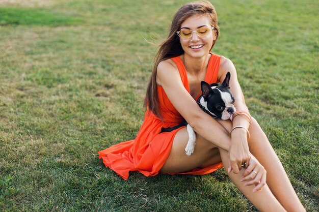 Portrait of happy pretty woman sitting on grass in summer park, holding boston terrier dog, smiling positive mood, wearing orange dress, trendy style, sunglasses, playing with pet