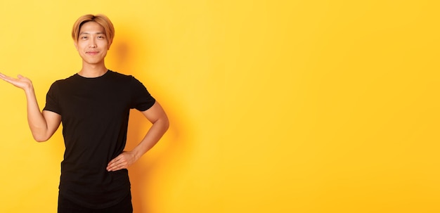 Portrait of happy and proud smiling asian guy holding something on hand over yellow background