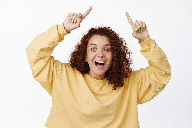 Portrait of happy redhead curly woman pointing fingers up, smiling on white