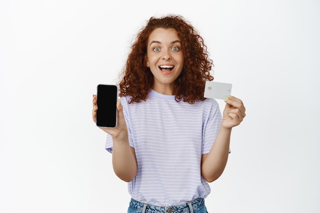 幸せな赤毛の女の子の肖像画は、携帯電話の画面、割引クレジットカード、驚いて笑顔、販売やアプリのプロモーション、白い背景について語っています。