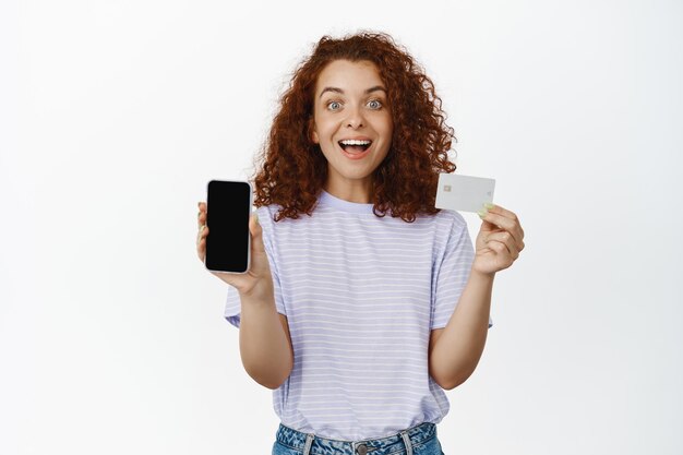 Portrait of happy redhead girl shows mobile phone screen, discount credit card, smiling amazed, telling about sale or app promo, white background.