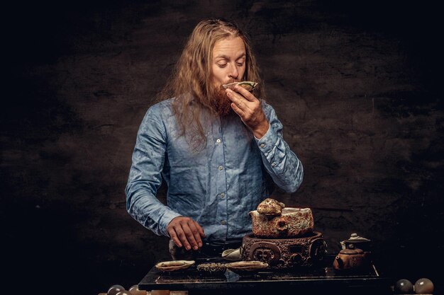 Portrait of a happy redhead hipster male with long hair and full beard dressed in a blue shirt, drinking hot tea from a small saucer. Isolated on a dark textured background.