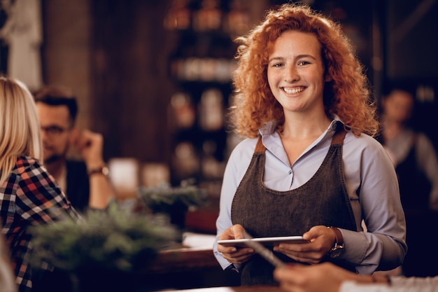 Portrait of happy redhead waitress holding touchpad while standing in a pub and looking at camera