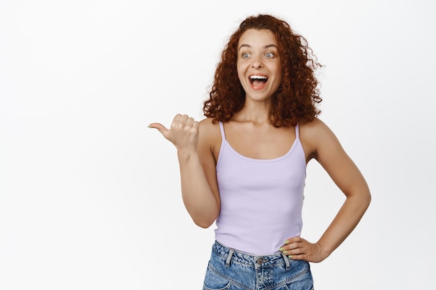 Portrait of happy redhead woman, ginger girl laughing, smiling and pointing left at awesome news, big sale promo, advertising smth, wearing summer clothes, white background.