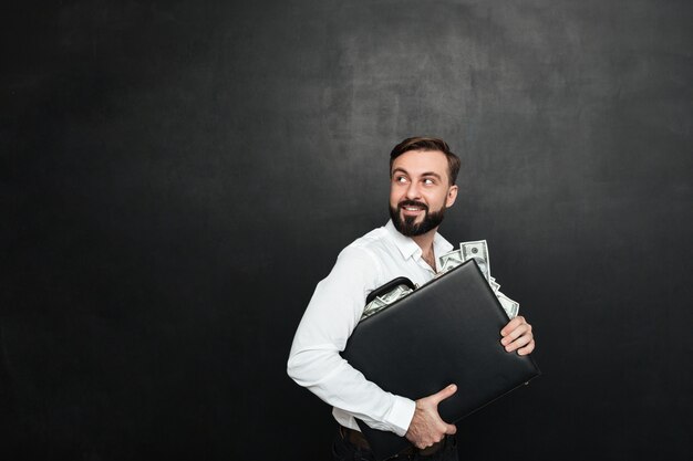 Portrait of happy rich businessman carrying briefcase full of dollar bills and looking back, isolated over dark gray