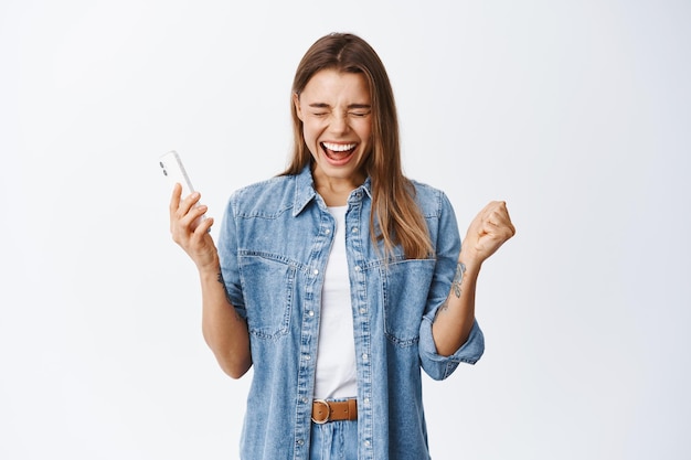 Portrait of happy screaming woman winning on smartphone, holding mobile phone and cheering, celebrating victory or achievement in internet, white