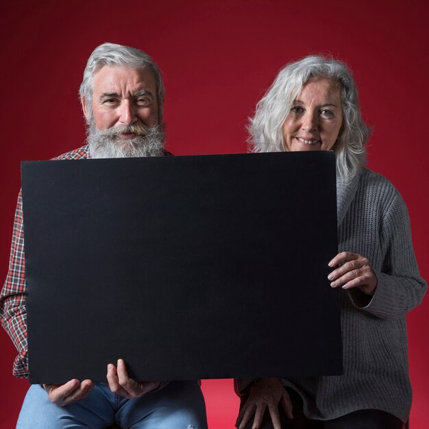 Portrait of happy senior couple holding black placard looking to camera against red background