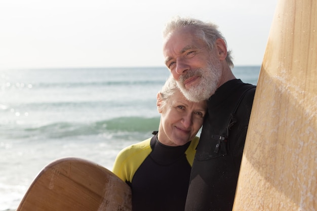 Portrait of happy senior couple with surfboards near sea. Grey-haired man and woman hugging, looking at camera and woman leaning mans chest. Relations, love and active rest of aged people concept