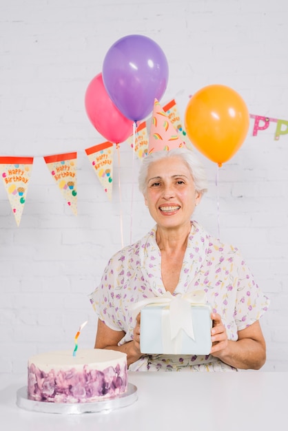 Portrait of a happy senior woman holding birthday gift with cake on desk