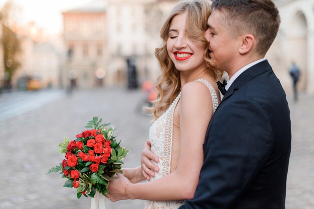 Portrait of happy smiled couple with bouquet made of red roses outdoors with closed eyes, romantic date