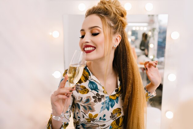 Портрет счастливой улыбающейся молодой женщины с роскошной прической, пьющей бокал шампанского в парикмахерской