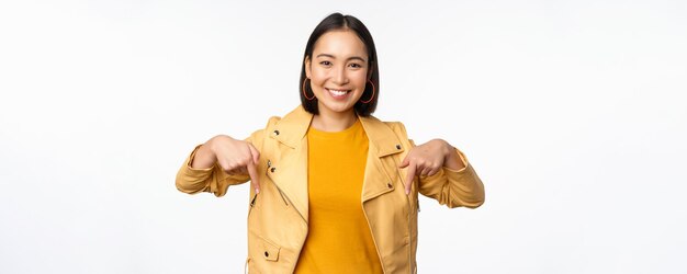 Портрет счастливой улыбающейся азиатской девушки, указывающей пальцем вниз и показывающей логотип, демонстрирующий баннер, стоящий в желтой куртке на белом фоне
