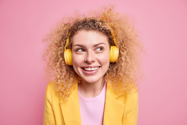 행복한 미소를 짓고 있는 곱슬머리 여성의 초상화는 무선 헤드폰을 통해 음악을 듣는 가장 좋아하는 재생 목록을 즐깁니다.