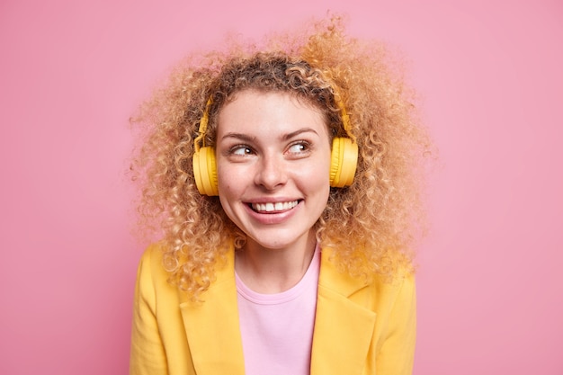 Portrait of happy smiling curly woman enjoys favorite playlist listens music via wireless headphones looks away grins at camera