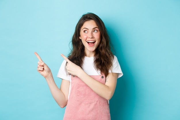 Портрет счастливой улыбающейся женщины-модели с гламурным румянцем, указывающей пальцами влево и смотрящей на промо-предложение, показывая пространство для рекламной копии, синий фон