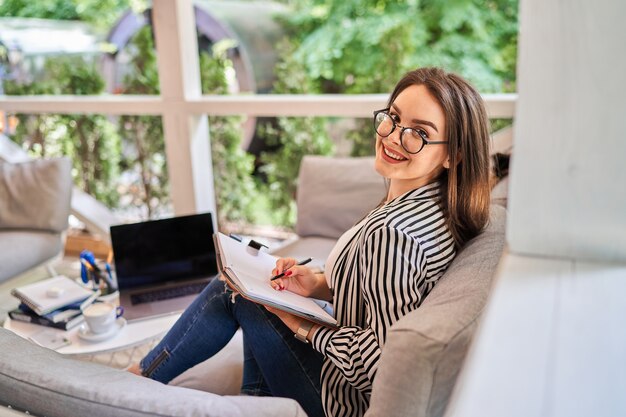 Portrait of happy smiling freelancer woman at home with notebook on the sofa.