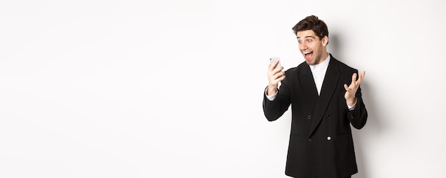 Free photo portrait of happy smiling man in black suit looking at smartphone screen with relieved and cheerful