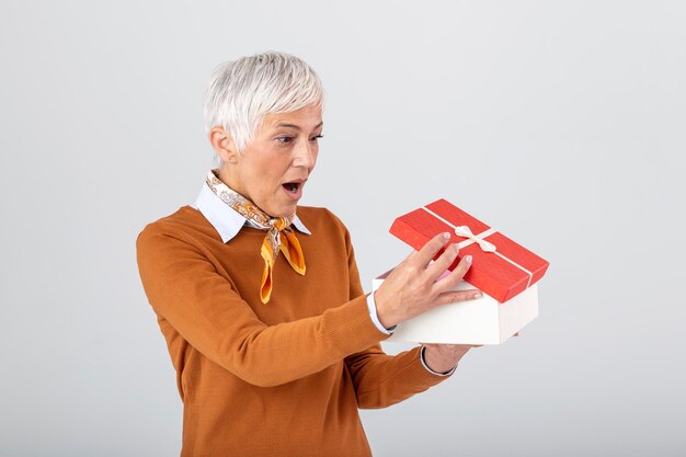 Portrait of a happy smiling mature woman opening a gift box isolated over gray background Excited senior casual woman holding present box