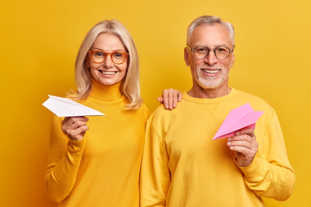 Portrait of happy smiling middle aged woman and man stand next to each other believe in good future hold papermade planes wear spectacles for good vision express positive emotions isolated on yellow
