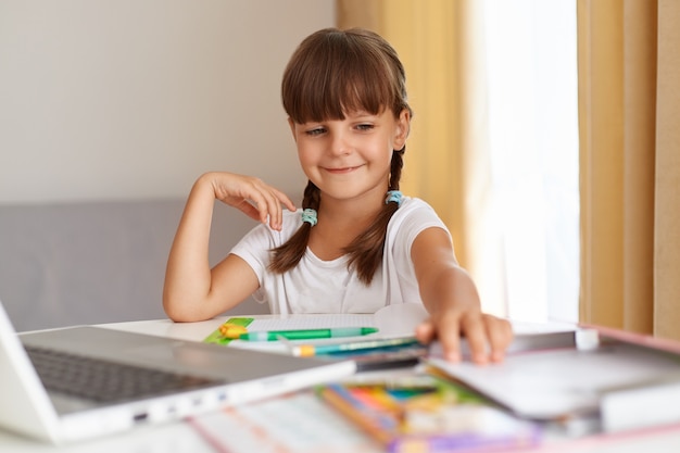 Портрет счастливого улыбающегося школьника в белой футболке, сидящего за столом против окна с занавесками перед ноутбуком, с позитивным выражением лица и выполняющего домашнее задание.