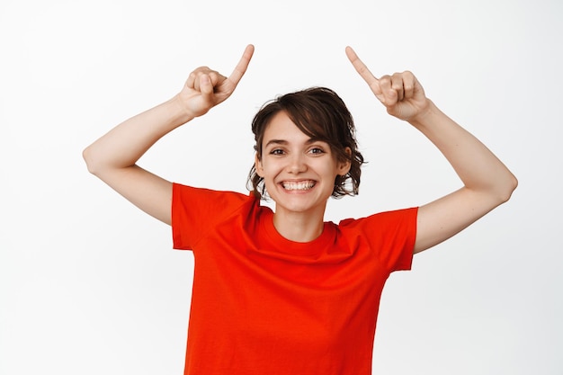 Portrait of happy smiling woman pointing fingers up, showing advertisement, give info, recommending website, white background.