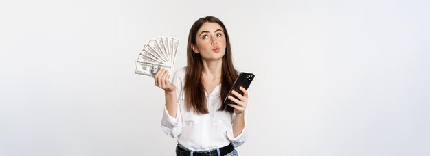 Free photo portrait of happy smiling woman using mobile phone app holding money cash standing over white backgr