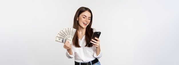 Portrait of happy smiling woman using mobile phone app holding money cash standing over white backgr