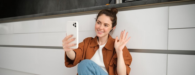 Free photo portrait of happy smiling woman video chats sits on kitchen floor at home talking to friend on