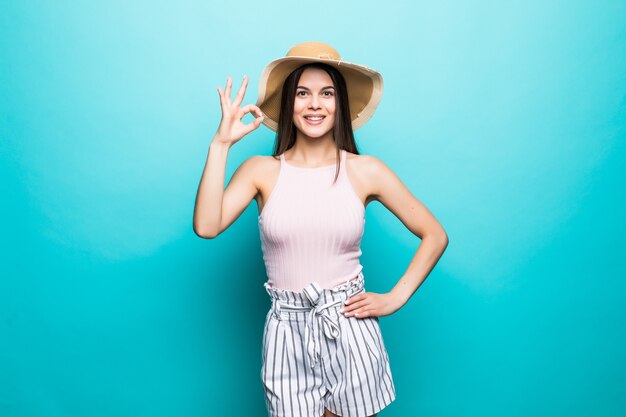 Portrait of happy smiling woman wearing dress, straw summer hat showing OK gesture, thumbs signal copy space isolated on blue wall.. People sincere emotions, lifestyle concept. Advertising area