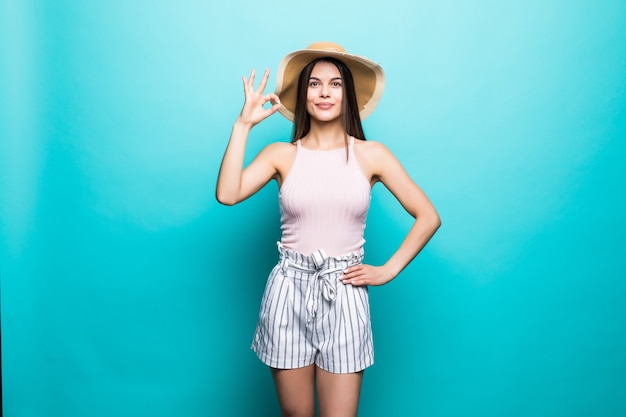 Portrait of happy smiling woman wearing dress, straw summer hat showing OK gesture, thumbs signal copy space isolated on blue wall.. People sincere emotions, lifestyle concept. Advertising area