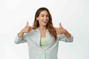 Free photo portrait of happy smiling woman winking, showing thumbs up in approval, like and agree, praise smth good, complimenting you, hinting on smth, standing over white background