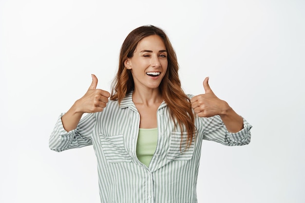 Portrait of happy smiling woman winking, showing thumbs up in approval, like and agree, praise smth good, complimenting you, hinting on smth, standing over white background