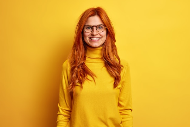Portrait of happy smiling woman with red hair stays always positive enjoys funny talk with friend dressed in turtleneck and spectacles.