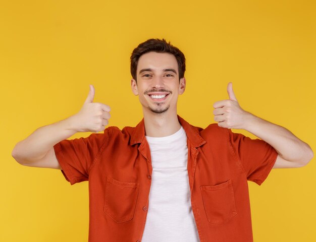 Portrait of happy smiling young man showing thumbs up gesture and looking at camera on isolated over yellow background