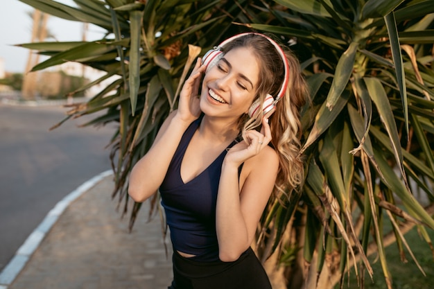 Portrait happy sunny morning of sportive young woman smiling with closed eyes, hearing music through headphones on street of tropical city. Training, workout, healthy lifestyle, cheerful mood
