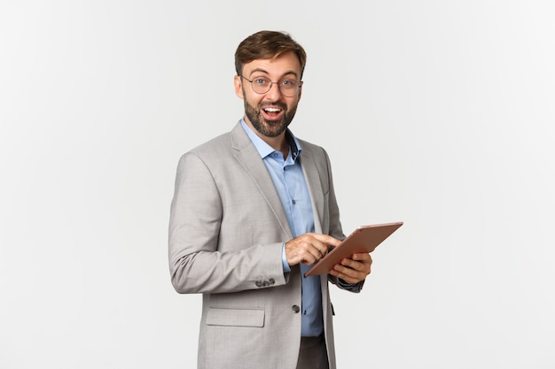 Портрет счастливого и удивленного бородатого бизнесмена в сером костюме и очках, держащего цифровой планшет ...