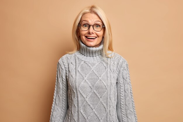 유럽의 모습으로 행복 놀란 금발 수석 여자의 초상화는 기꺼이 안경을 착용하고 따뜻한 회색 스웨터는 대담 자로부터 즐거운 소식을 듣고 경이로움을 표현합니다.