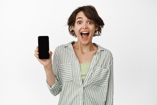 Portrait of happy and surprised cute girl showing her phone screen and looking in awe, recommending download an app, standing on white.