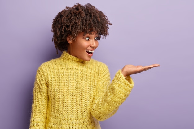Portrait of happy surprised woman with dark skin, curly hairstyle, raises palm over blank space, sees something awesome, holds invisible object, isolated over purple wall. Wow, it is great