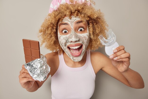 Portrait of happy surprised young woman stares at camera holds bar of chocolate and mouth expander has amazed expression applies beauty mask on face for skin treatment wears bathhat and t shirt