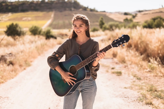 Ritratto di un adolescente felice che sta sulla pista non asfaltata che gioca chitarra