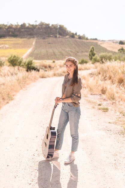 Портрет счастливой девушки-подростка с гитарой, стоящей на грунтовой дороге