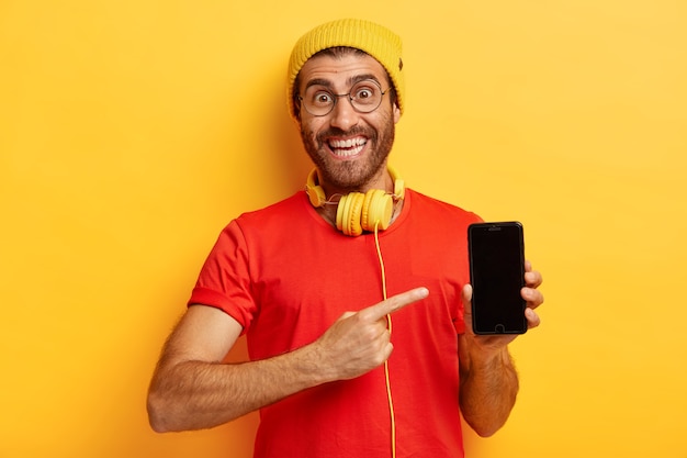 행복하지 않은 남자의 초상화가 스마트 폰 화면에서 가리키고 디스플레이를 보여주고 새 전자 장치를 구입하고 세련된 모자와 캐주얼 빨간색 티셔츠를 입고 노란색 벽에 모델을 착용합니다.