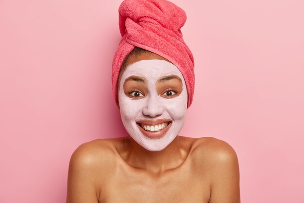 Portrait of happy woman applies clay nourishing facial mask, has glad expression, being in good mood, enjoys rejuvenation treatment, wears rosy towel on wet hair