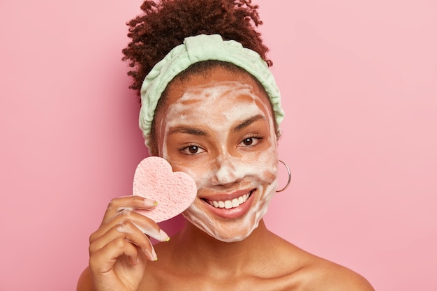 Portrait of happy woman has perfect well cared skin, applies foaming soap for washing face, has pleased expression, holds heart shaped sponge for wiping makeup