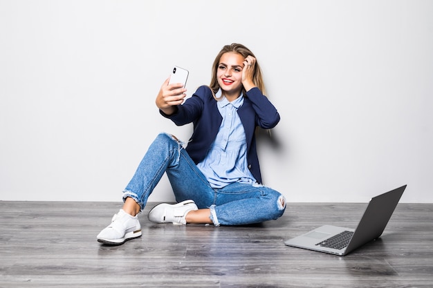 행복한 여자가 바닥에 앉아 회색 벽에 스마트 폰으로 셀카 사진을 만드는 초상화