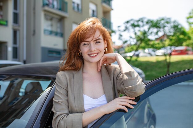 通りに車のそばに立っている幸せな女性の肖像画ドアを開けた車の後ろに立っている若いかわいいコーカサインの女性
