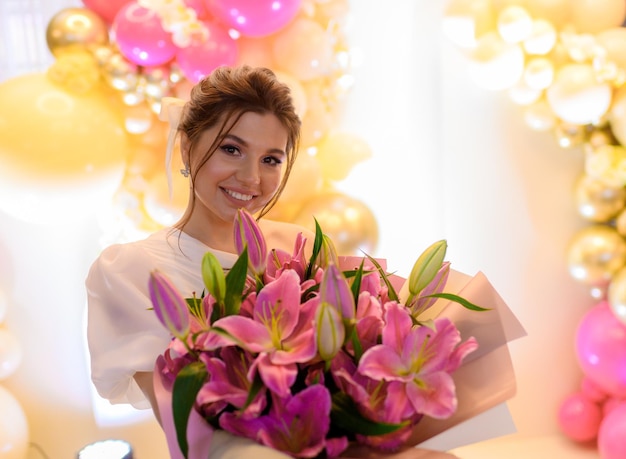 Portrait of happy woman with brunette hairdo dressed in trendy gown holding large flower arrangement with pink lilies smiling and looking at camera during wedding celebration in restaurant