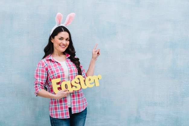 Portrait of a happy woman with bunny ears on head holding easter word pointing finger upward on blue background