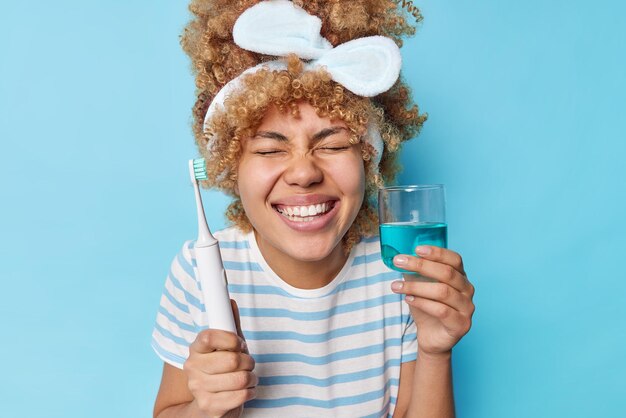 巻き毛の櫛の髪を持つ幸せな女性の肖像画はカジュアルなストライプのTシャツを着ており、ヘッドバンドは歯ブラシを保持し、新鮮なうがい薬は青い背景の上に定期的に隔離された歯をきれいにします口腔衛生